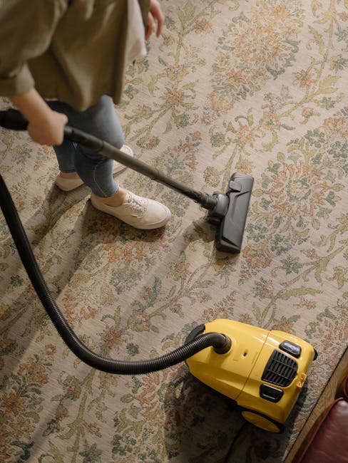 A professional cleaner dressed in a full white protective suit, face mask, and blue gloves is performing deep cleaning or sanitisation of a beige carpet in a well-lit living room. The cleaner is using