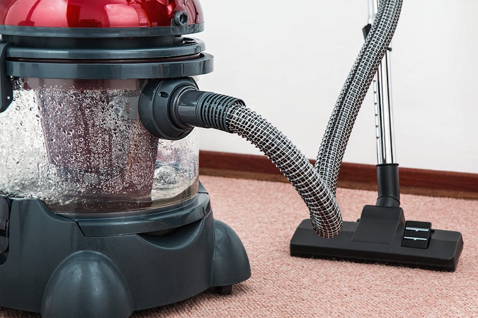 A person is performing surface cleaning on a patterned area rug with a vacuum cleaner in a domestic setting. The individual, wearing beige shoes and casual clothing, is holding a black vacuum hose att